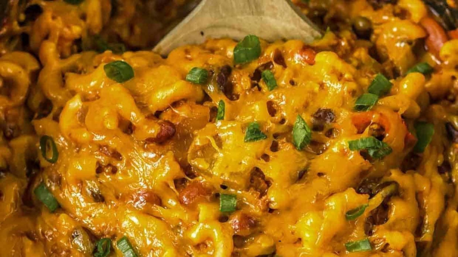 Close-up of cheesy macaroni casserole with melted cheese, ground meat, beans, and chopped green onions, being stirred with a wooden spoon.