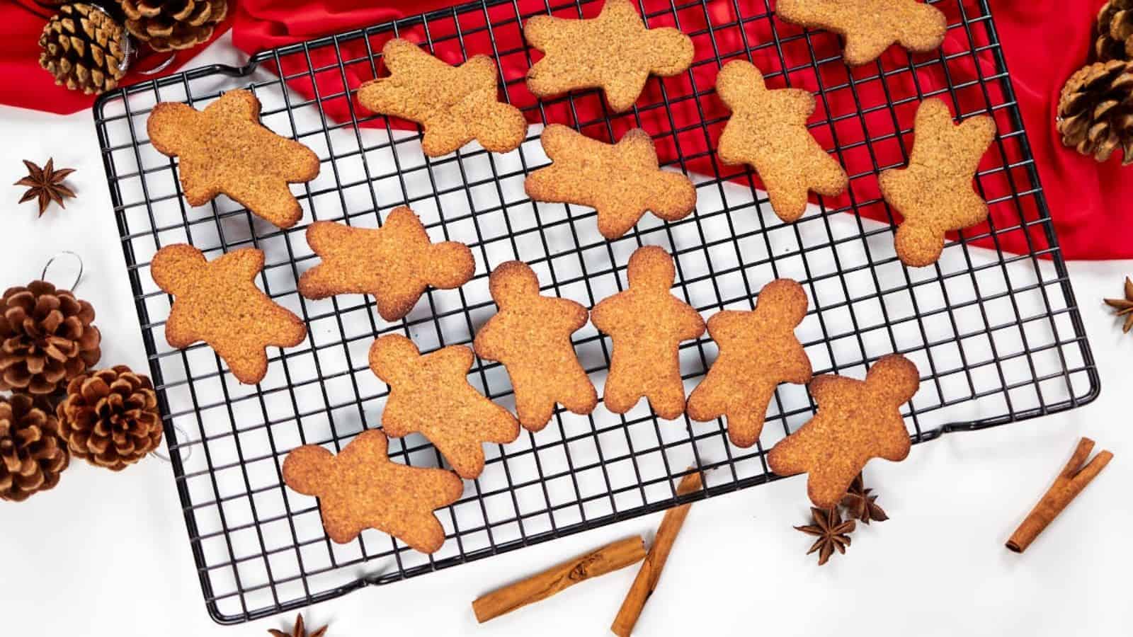 Gingerbread cookies in the shape of people cooling on a wire rack, with cinnamon sticks, pine cones, and red fabric nearby.