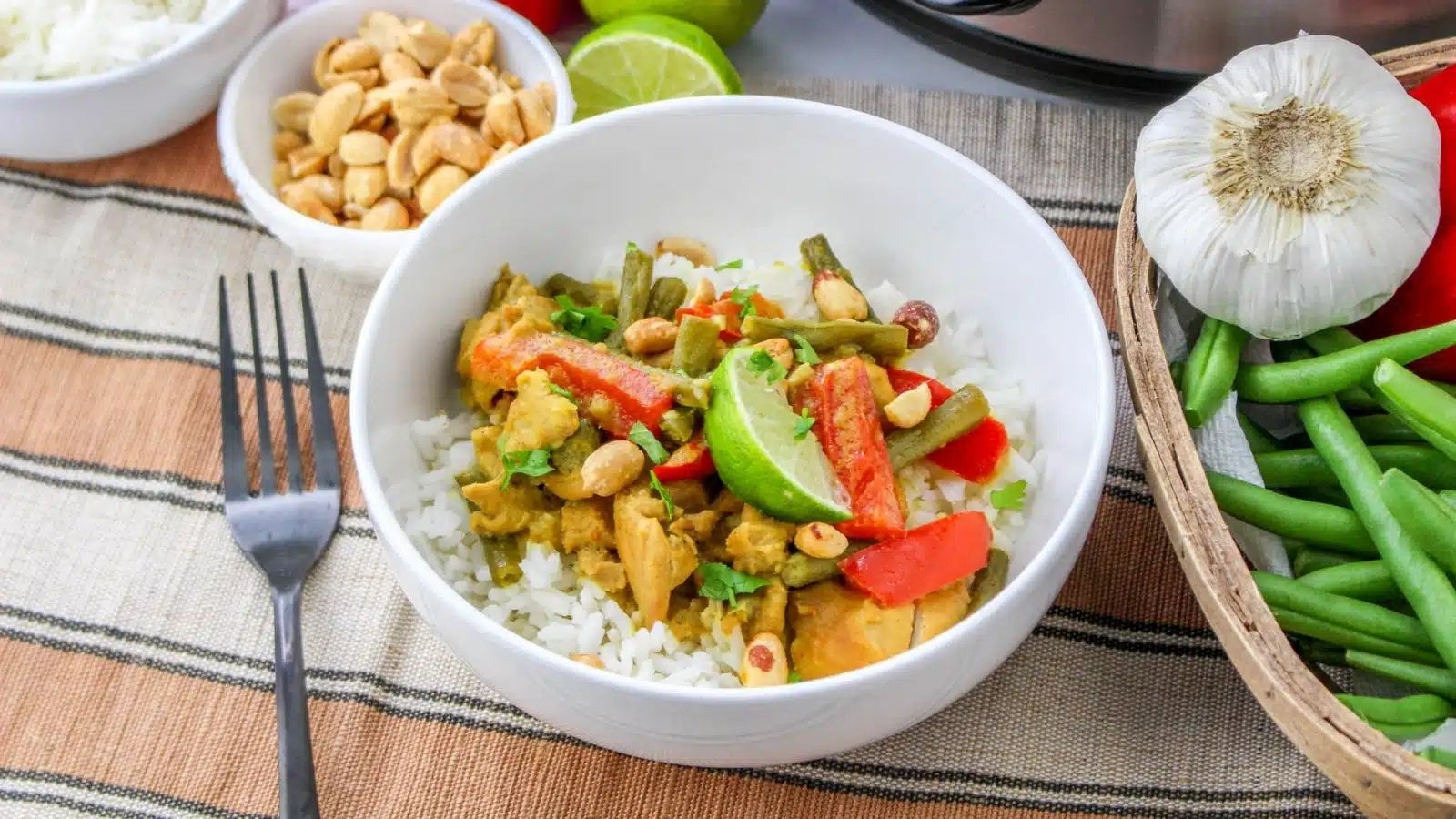 A bowl of rice topped with curry chicken, red peppers, green beans, and peanuts, garnished with lime wedges, next to a fork. A basket with vegetables and a bowl of peanuts beside it.