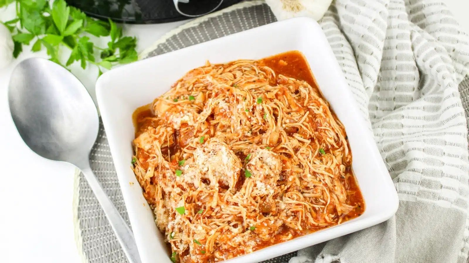 A square white bowl filled with shredded chicken in tomato sauce, garnished with herbs, next to a silver spoon and a striped napkin.