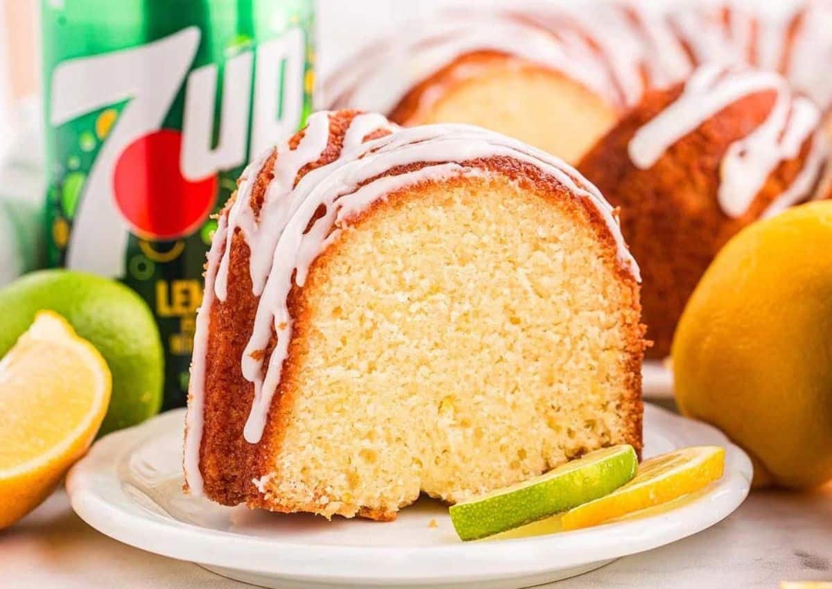 A slice of bundt cake with white icing is served on a plate with lemon and lime wedges; a 7UP soda bottle is in the background.