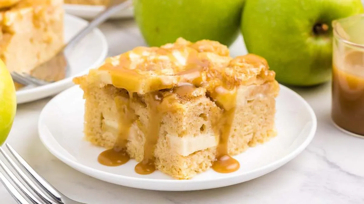 A slice of apple cake topped with caramel sauce is served on a white plate, with green apples and a glass of caramel in the background.