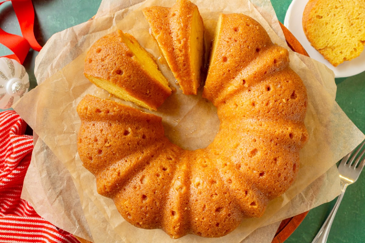 A golden-brown Bundt cake with several slices cut, displayed on parchment paper, next to a plate holding a slice, a fork, and a red-striped cloth.