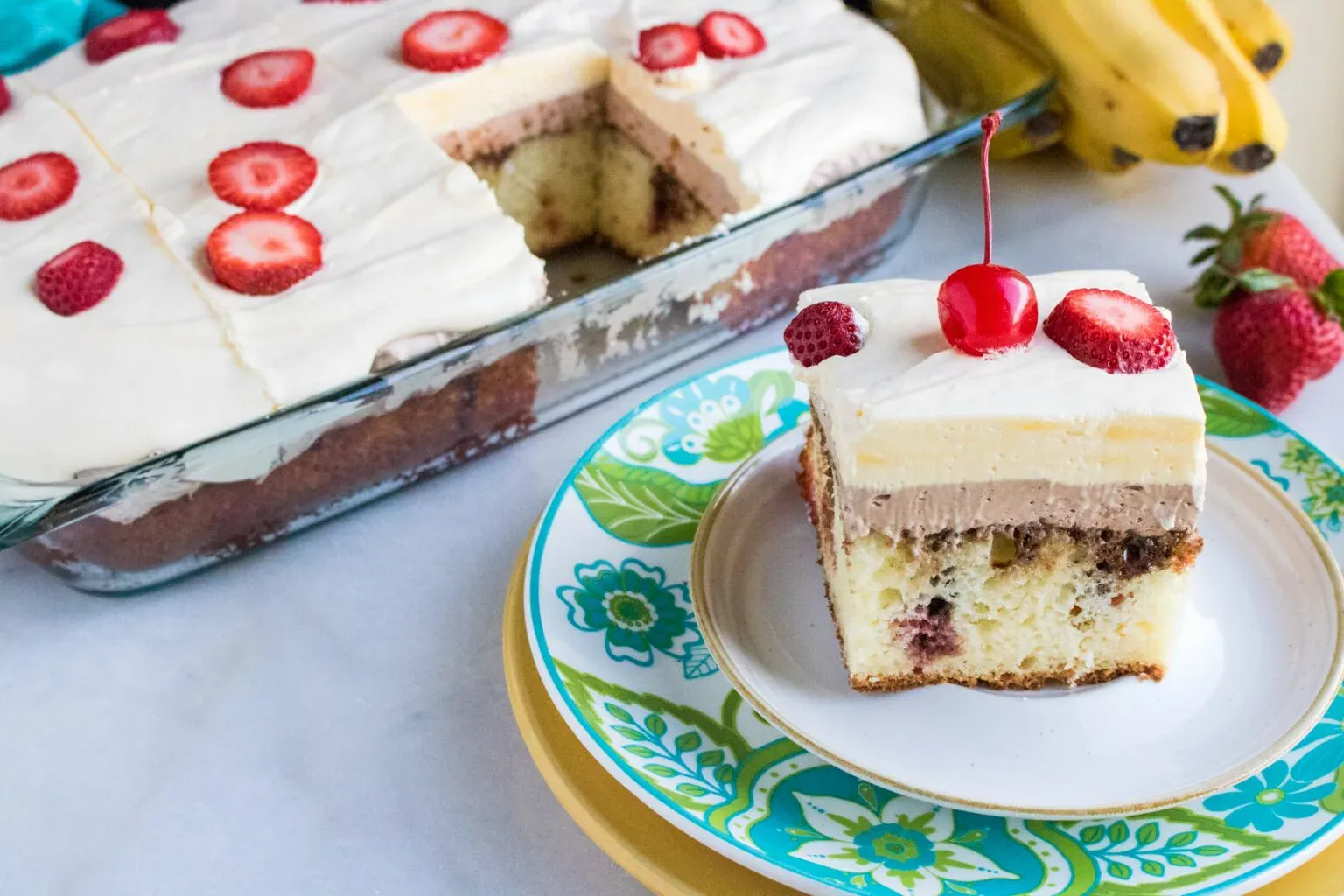 A slice of layered cake with whipped topping, strawberries, and a cherry sits on a plate, with the rest of the cake in a glass pan nearby. Bananas and strawberries are in the background.
