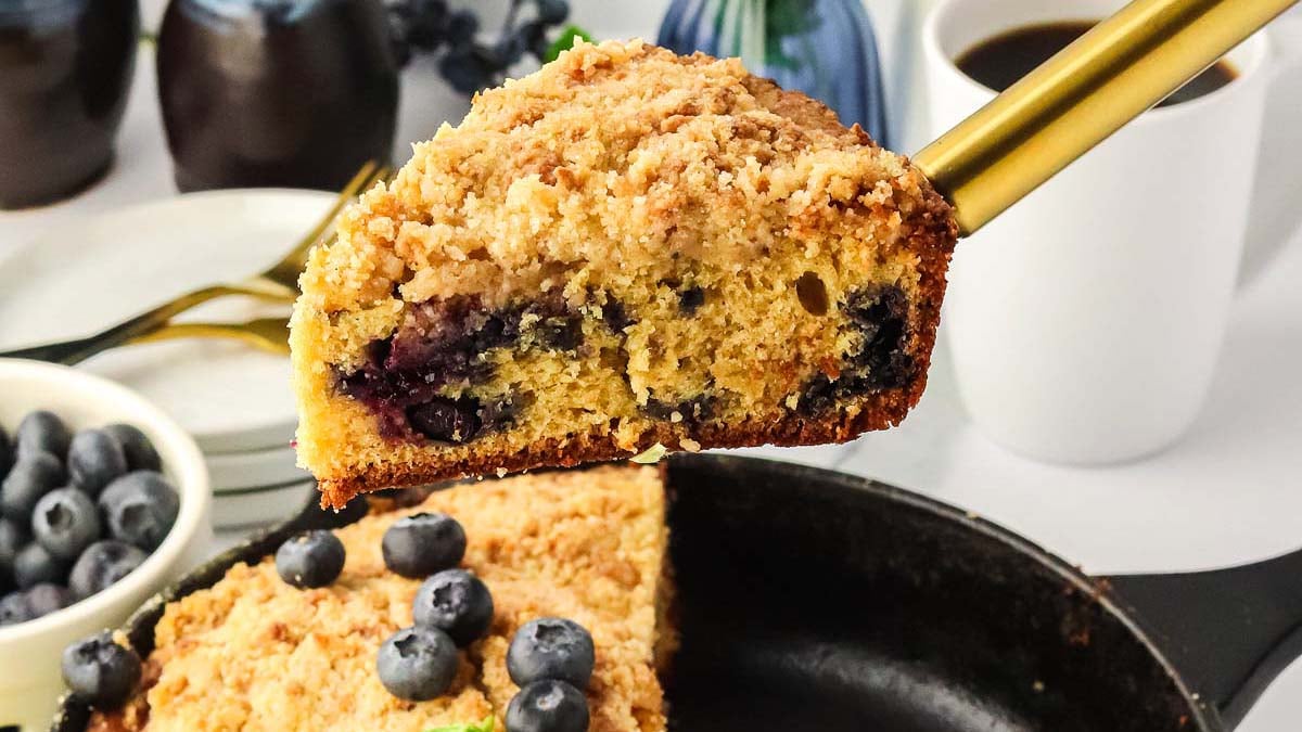 A slice of blueberry crumb cake on a spatula is held above a pan, with fresh blueberries, coffee, and plates in the background.