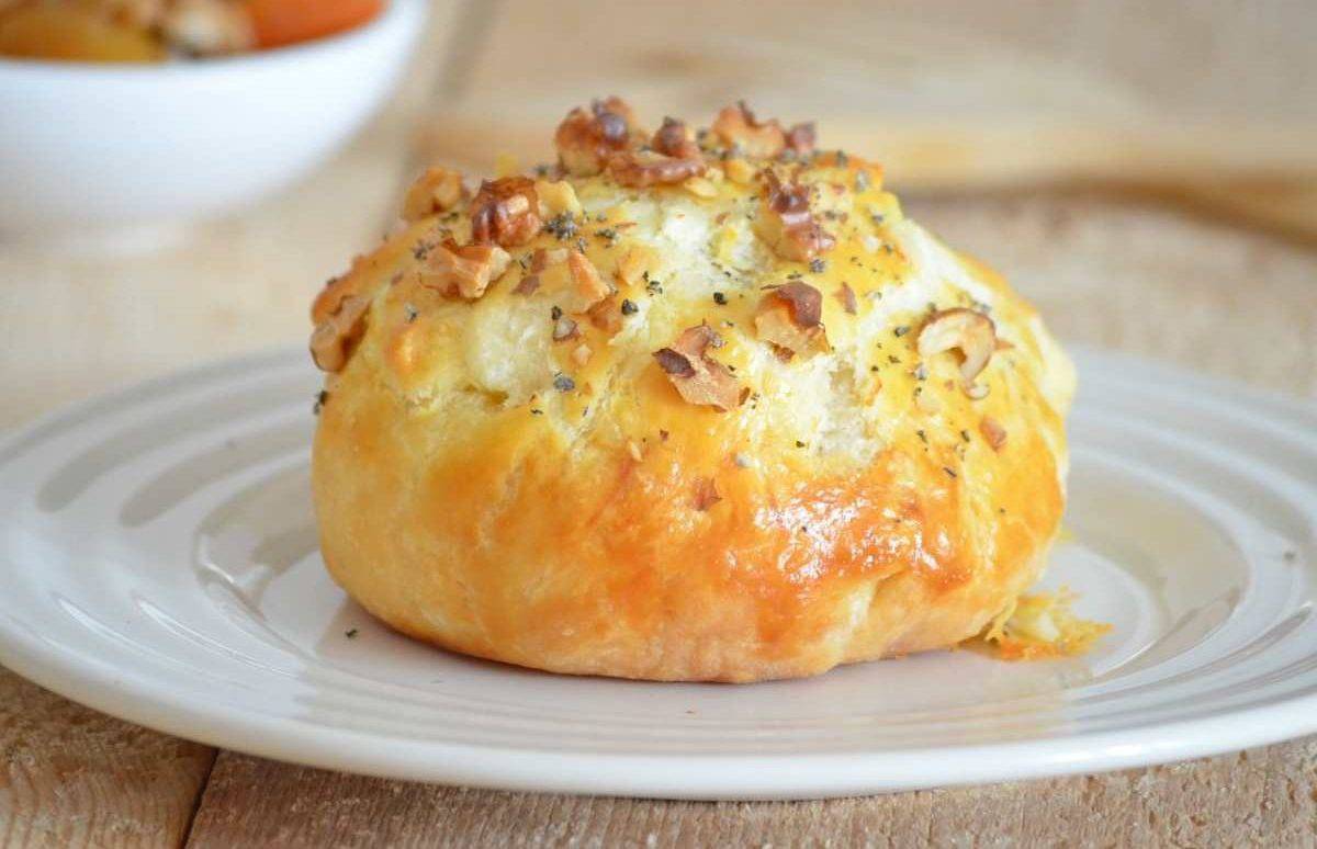 A baked pastry topped with chopped nuts and herbs sits on a white plate, with a bowl in the blurred background.