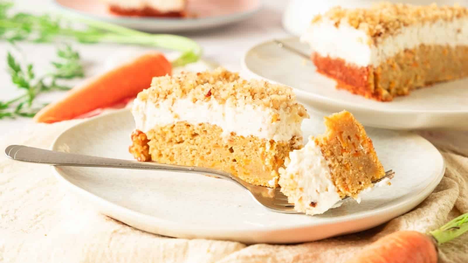 A slice of carrot cake with cream cheese frosting and crumb topping is served on a plate with a fork; whole carrots are visible in the background.