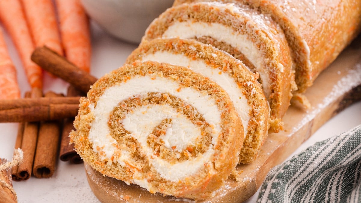 Sliced carrot cake roll with cream cheese filling on a wooden board, with cinnamon sticks and whole carrots in the background.