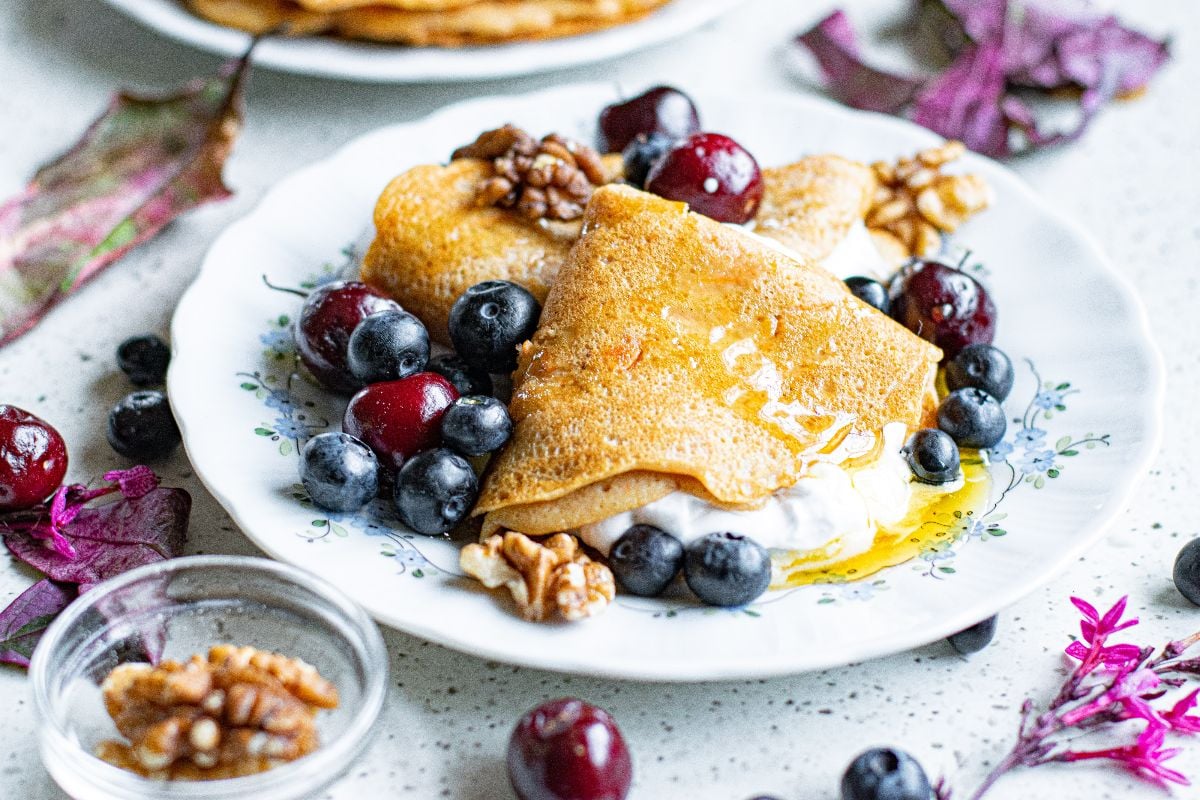 Plate with folded crepes topped with blueberries, cherries, walnuts, honey, and a dollop of cream, surrounded by fresh fruit and leaves.