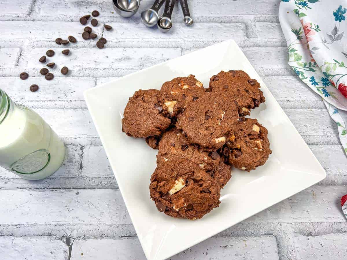 A square white plate holds a pile of chocolate cookies with visible chocolate chips. A bottle of milk, measuring spoons, and a floral napkin are nearby on a white brick surface.
