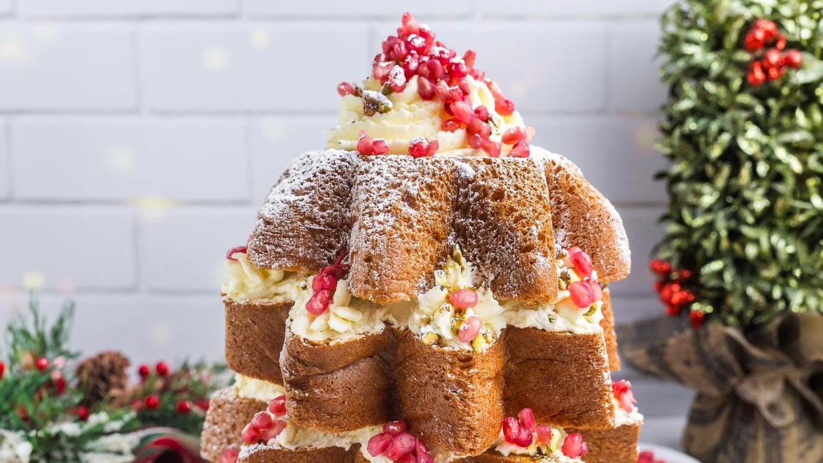 A layered star-shaped cake dusted with powdered sugar, filled with cream and topped with pomegranate seeds, sits on a festive table with holiday decorations in the background.