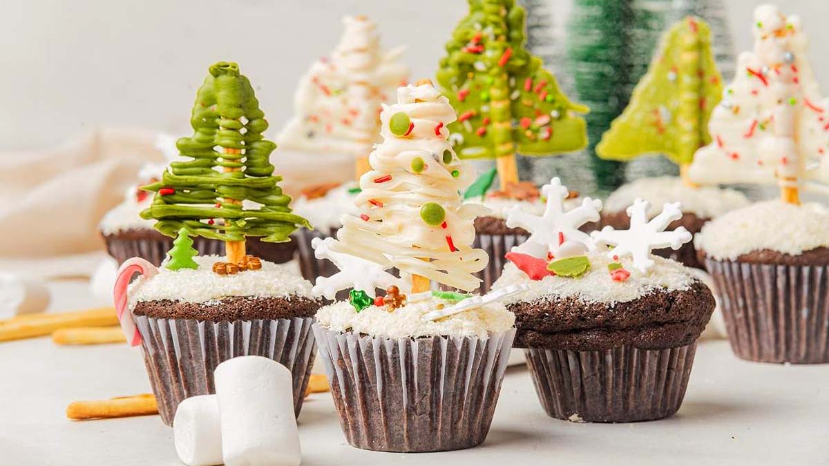Chocolate cupcakes decorated with frosting and candy to resemble Christmas trees and snowflakes, displayed on a white surface with marshmallows and pretzel sticks nearby.