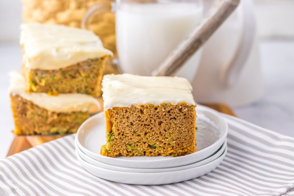 A slice of frosted zucchini cake on a stack of white plates, with more cake and a glass of milk in the background.