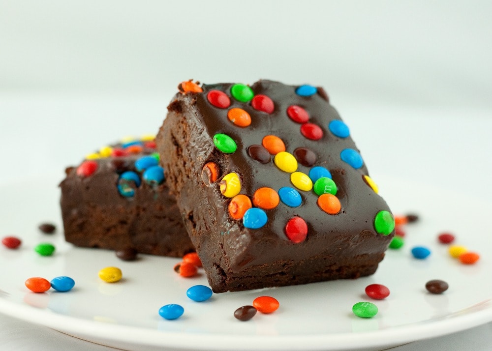 Two chocolate brownies topped with colorful candy-coated chocolates are stacked on a white plate, with a few candies scattered around.