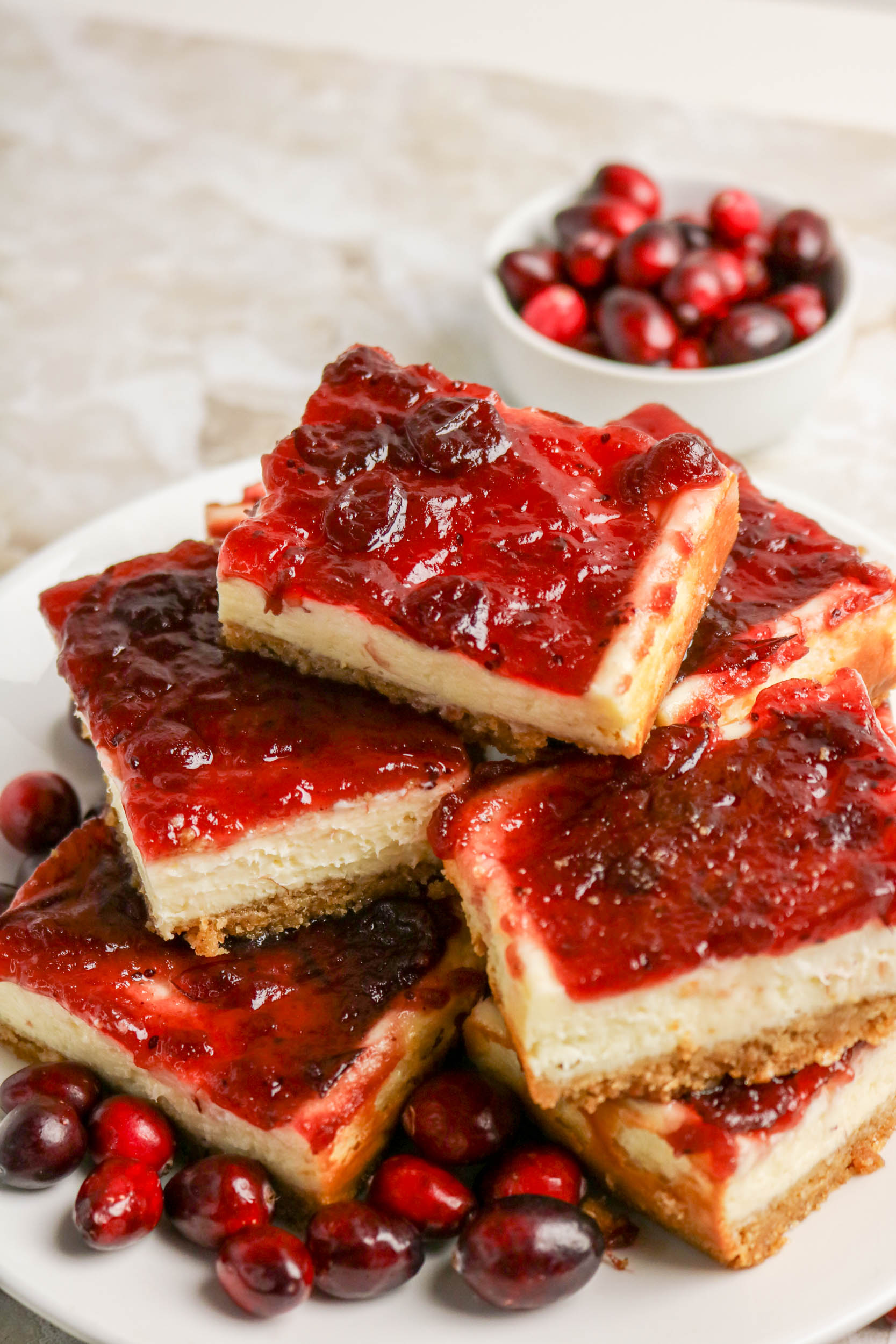A plate of cheesecake bars topped with cranberry sauce, surrounded by fresh cranberries, with a small bowl of cranberries in the background.
