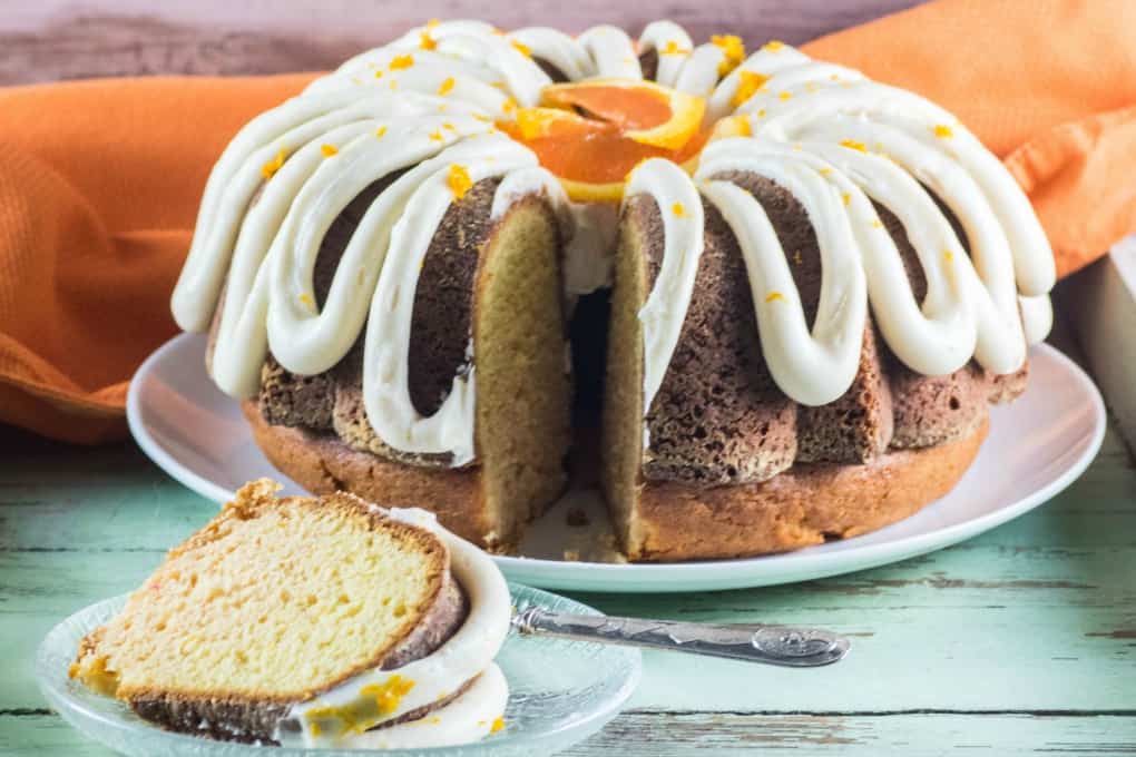 A bundt cake with white icing, garnished with an orange slice and zest, sits on a plate; a slice is served on a glass dish with a fork.