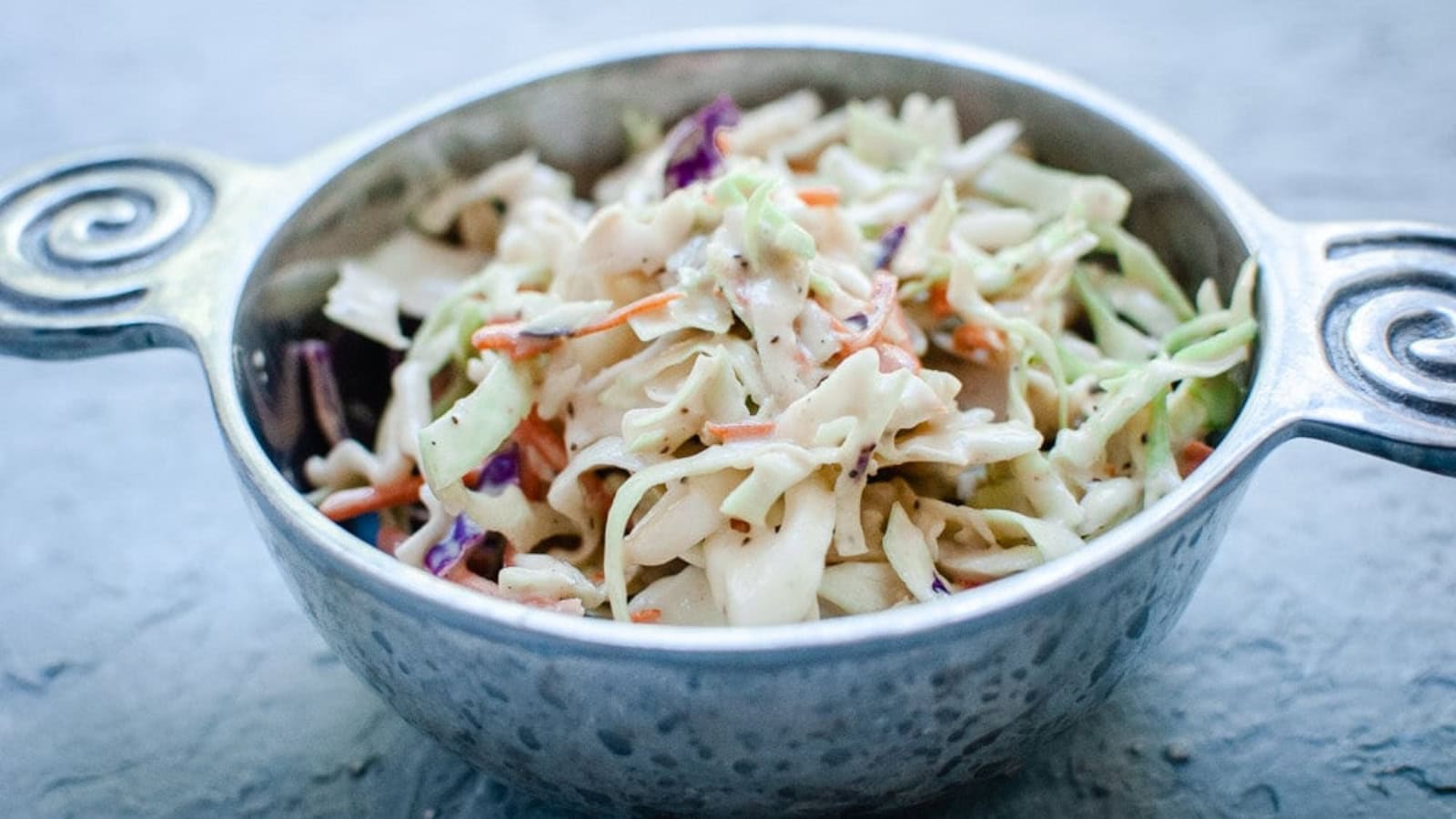 A metal bowl with handles contains creamy coleslaw made of shredded cabbage, carrots, and purple cabbage.