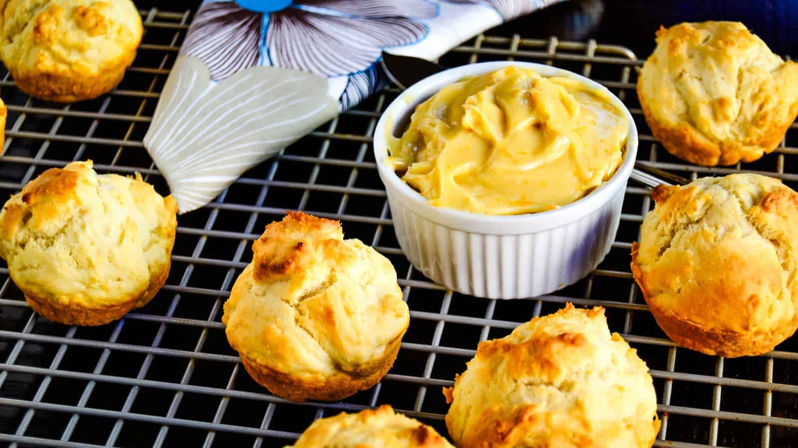 Golden brown drop biscuits on a cooling rack, arranged around a ramekin filled with whipped butter; a patterned napkin is in the background.