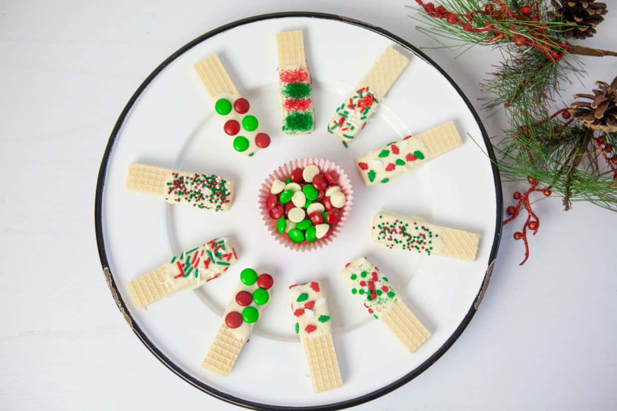 A white plate with wafer cookies dipped in white chocolate and decorated with colorful sprinkles, arranged in a circle around a cup of red, green, and white candy-coated chocolates.