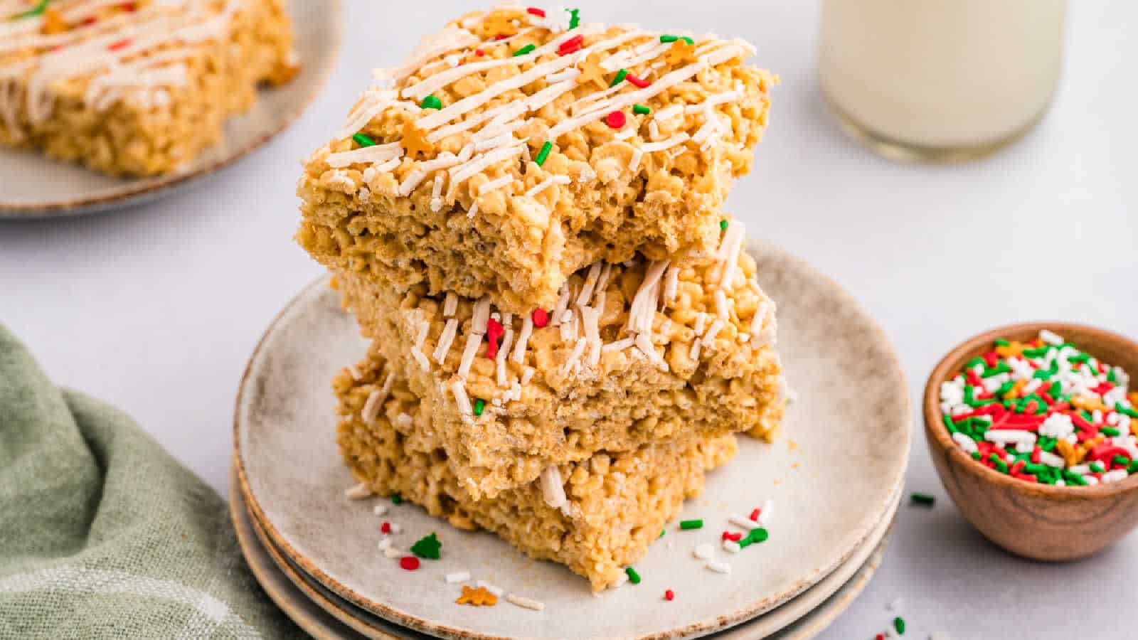 Two stacked rice crispy treats with white drizzle and red, green, and white sprinkles on a plate, with a bowl of sprinkles and glass of milk nearby.