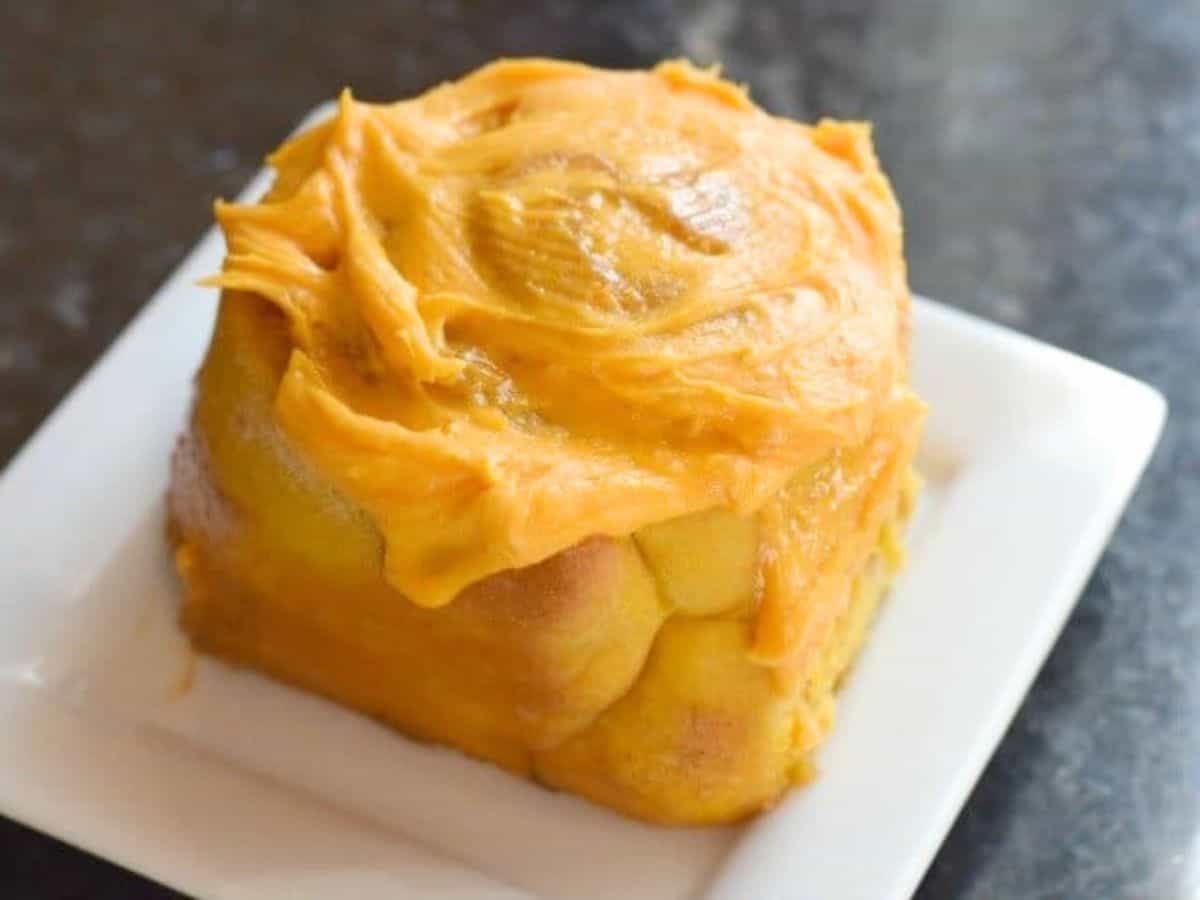 A single cinnamon roll topped with orange frosting sits on a white square plate against a dark background.