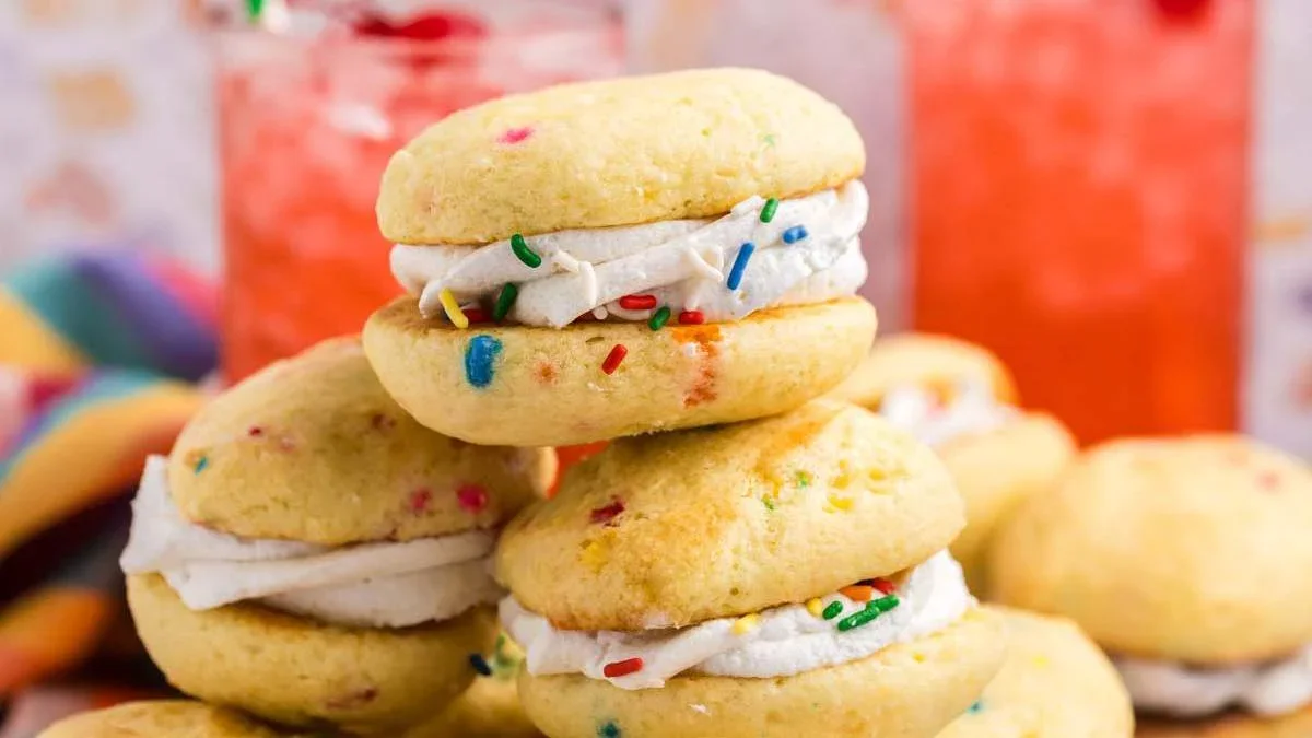 Several funfetti cookies are sandwiched with white frosting and topped with colorful sprinkles, stacked in front of two glasses with red drinks.