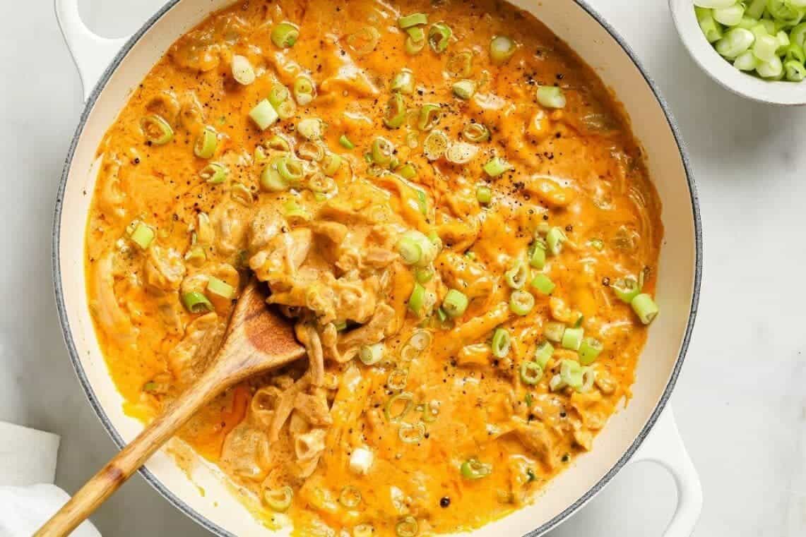 A creamy orange sauce with sliced green onions in a white pan, stirred with a wooden spoon.