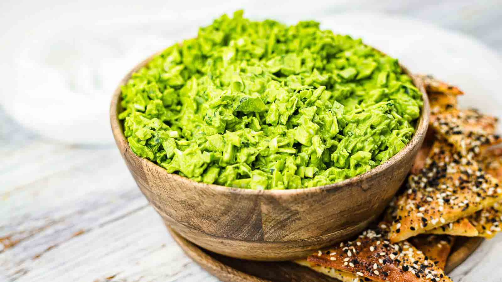 A wooden bowl filled with green chopped salad sits next to pieces of seeded flatbread on a round wooden tray.