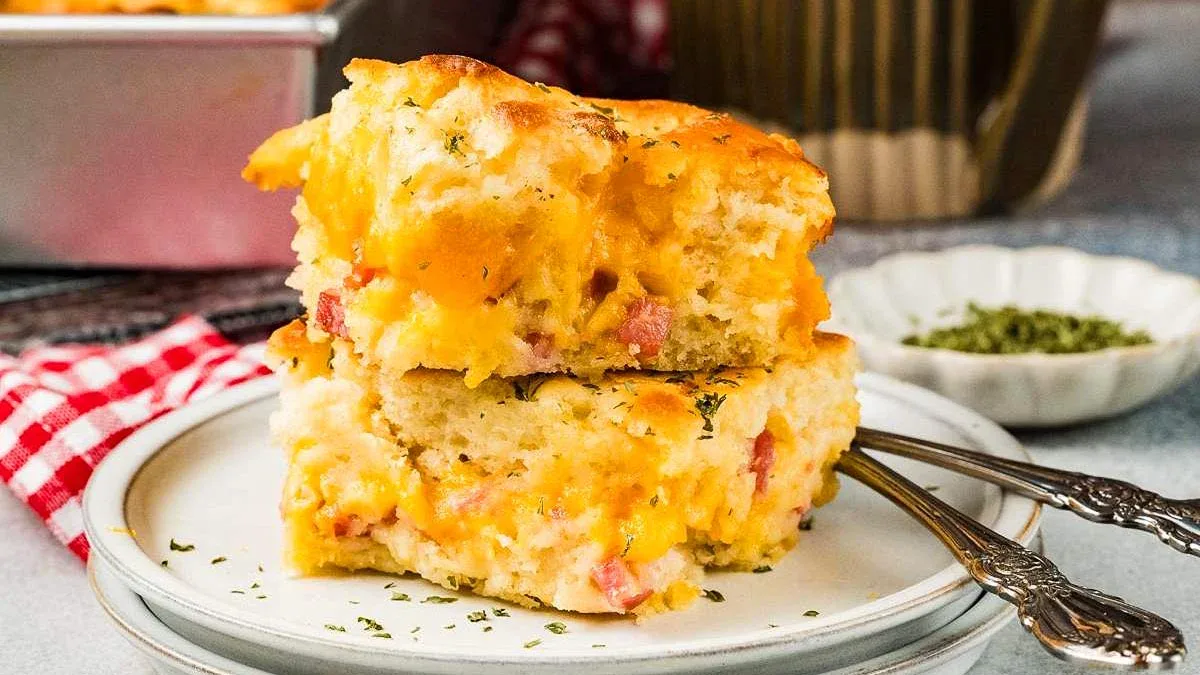 Two pieces of cheesy cornbread with bits of ham are stacked on a plate with two forks, garnished with herbs.