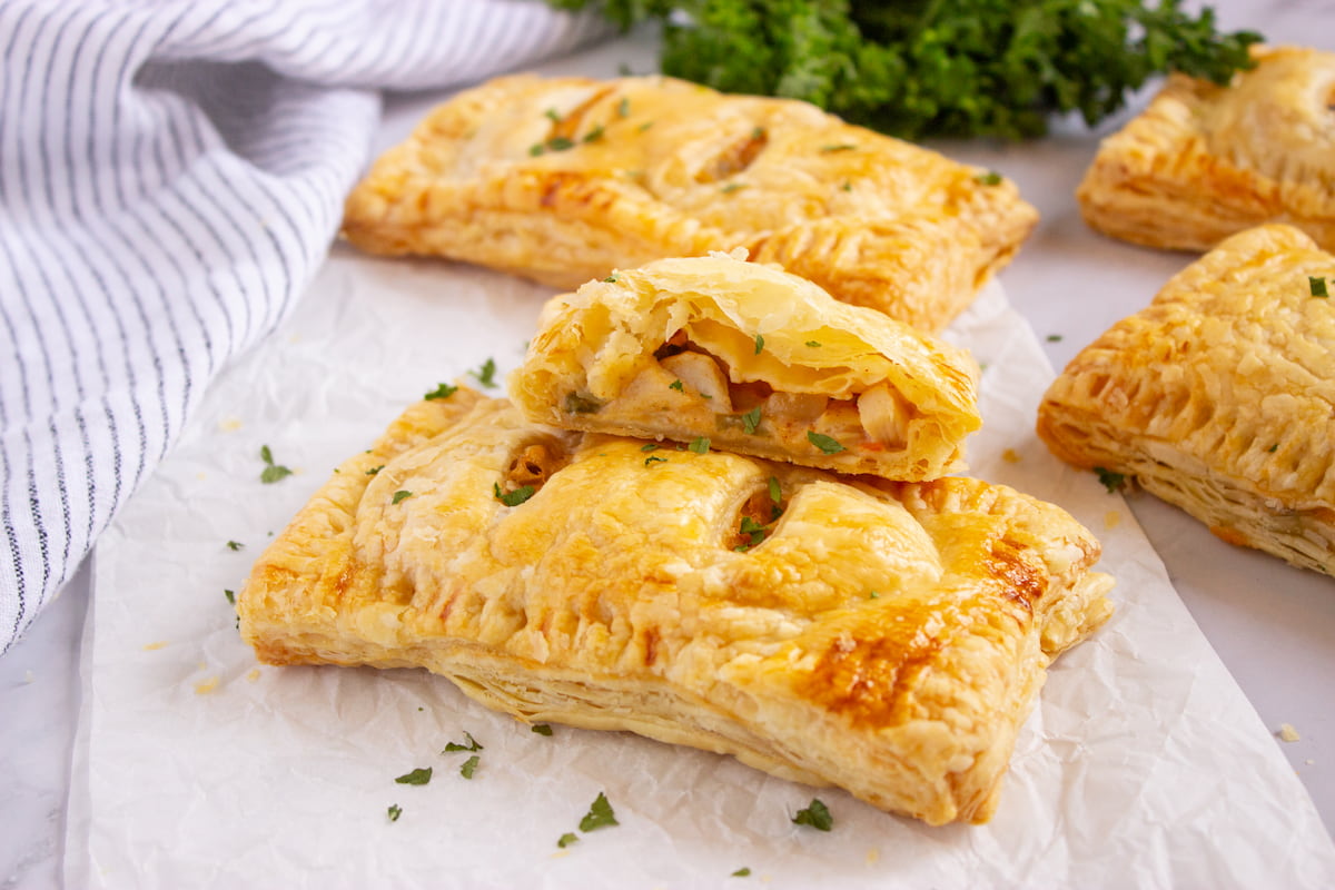 Golden puff pastry pockets, one cut open to show a filling of diced chicken and vegetables, arranged on parchment paper with parsley garnish in the background.