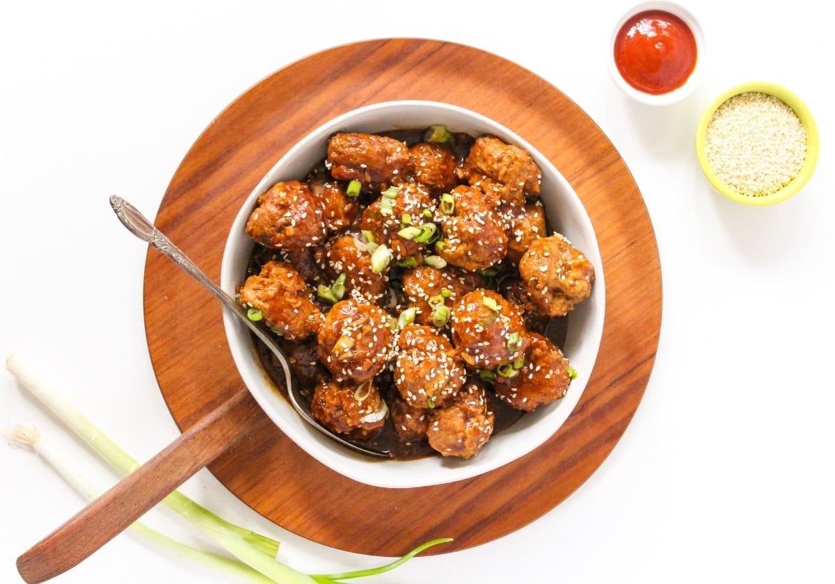 A white bowl of glazed meatballs garnished with sesame seeds and chopped green onions, placed on a wooden board with a spoon, spring onions, sesame seeds, and sauce on the side.