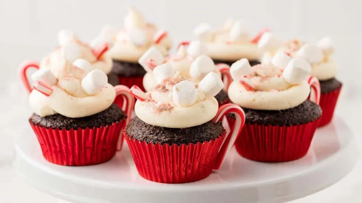 Chocolate cupcakes in red wrappers topped with white frosting, mini marshmallows, crushed candy, and small candy canes, arranged on a white cake stand.