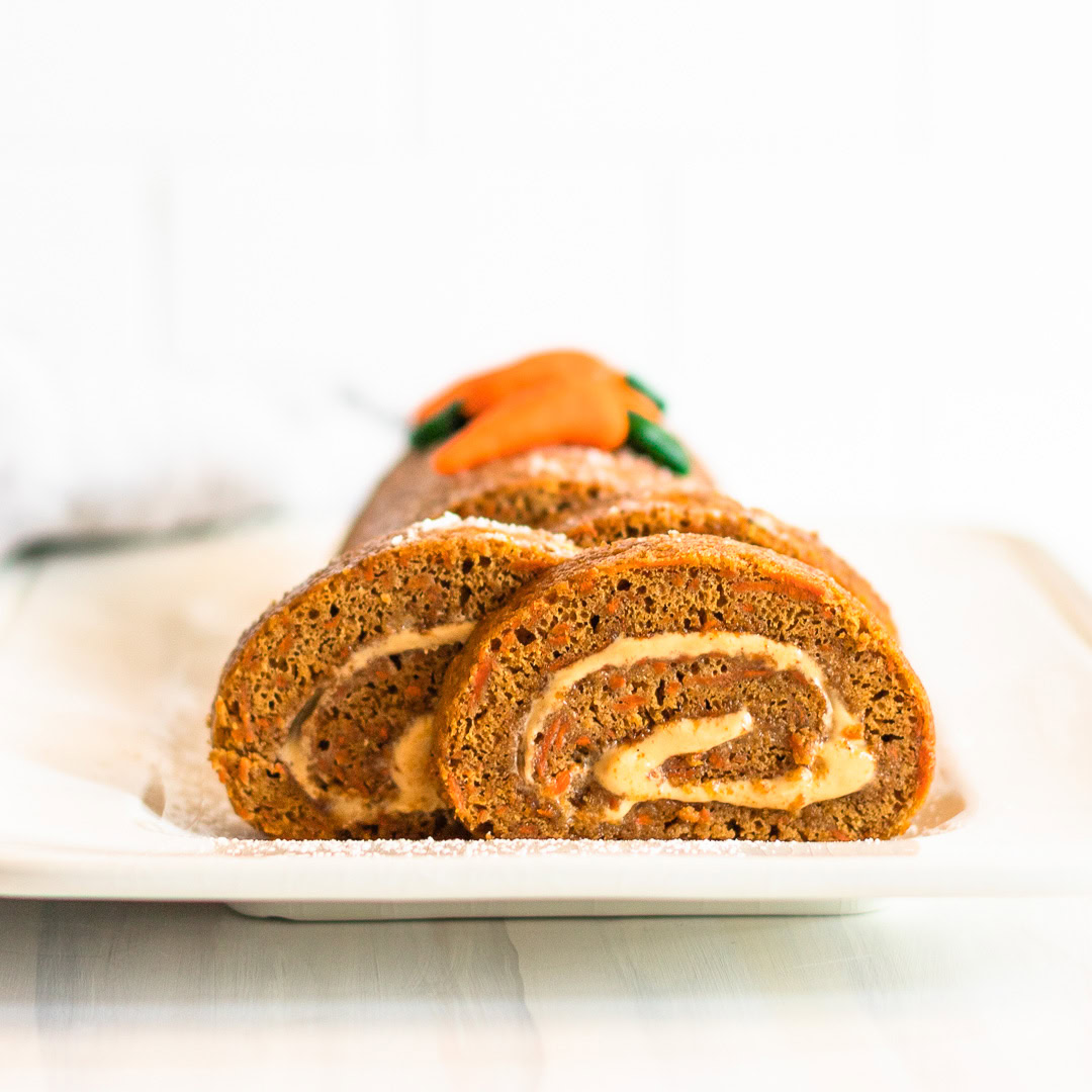 Two slices of carrot cake roll with cream cheese filling are served on a white rectangular plate, with the rest of the roll in the background.