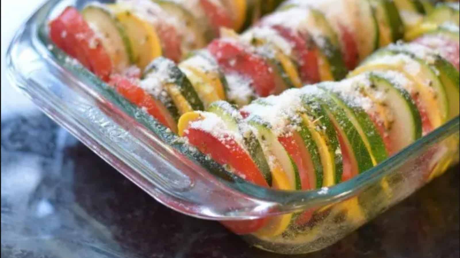 A glass baking dish filled with neatly arranged slices of zucchini, yellow squash, and tomato, topped with grated cheese or breadcrumbs, ready to be cooked.