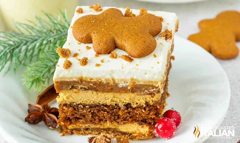 A slice of layered gingerbread dessert topped with a gingerbread cookie and crumbs, garnished with cranberries and pine on a white plate.