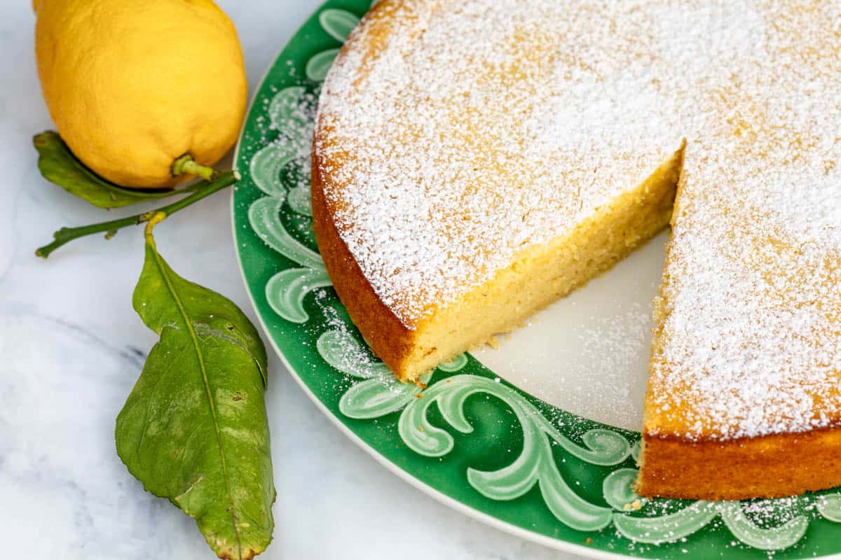 A round lemon cake dusted with powdered sugar sits on a green plate; one slice is cut. A whole lemon with leaves is next to the cake.