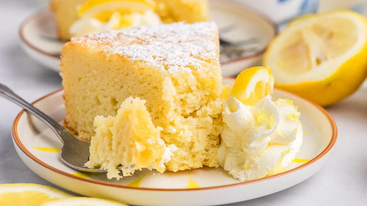 A slice of lemon cake with powdered sugar on top, served with whipped cream and a lemon slice on a plate, with a fork and a halved lemon in the background.