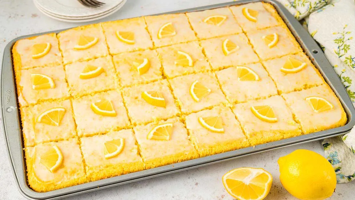 A rectangular baking tray with 24 lemon bars, each topped with a small lemon slice. Whole and cut lemons are placed beside the tray.