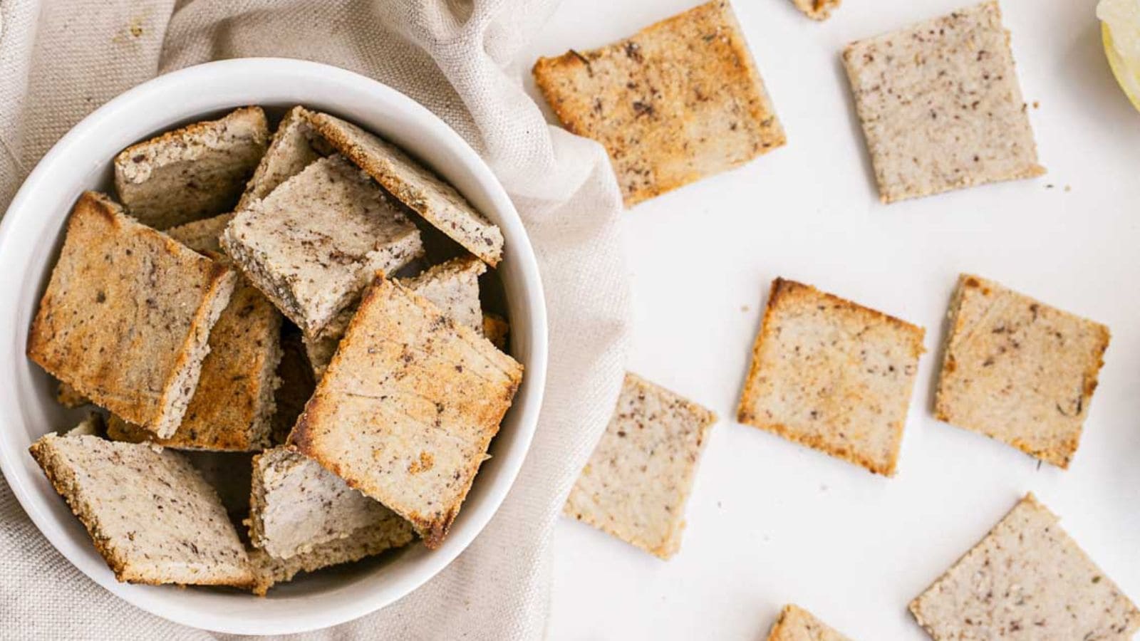 A white bowl filled with square, baked crackers sits on a light fabric, with several crackers scattered on a white surface nearby.