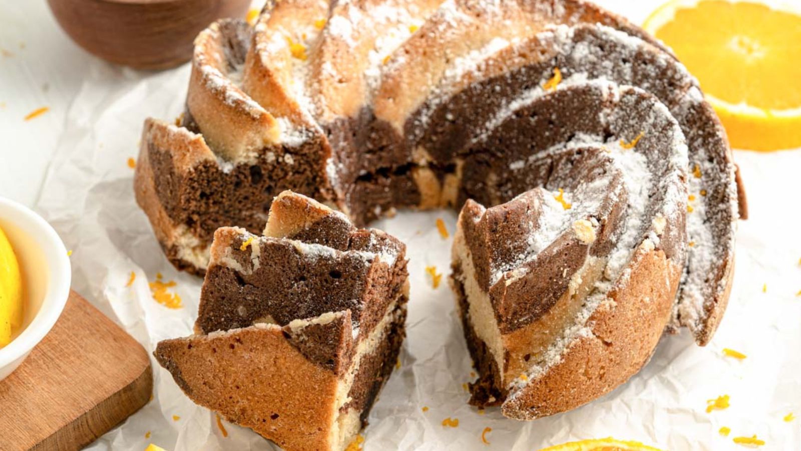 A marble bundt cake with a swirl pattern, sliced to show chocolate and vanilla layers, is displayed on parchment paper with orange slices nearby.