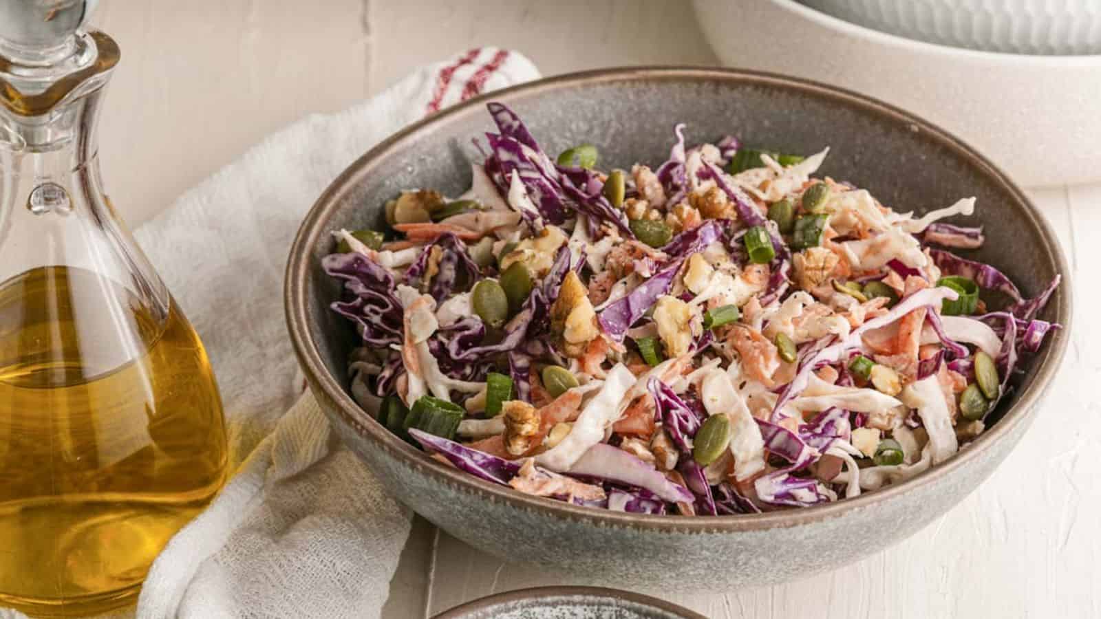 A bowl of coleslaw made with shredded red and green cabbage, seeds, nuts, and dressing sits on a table beside a bottle of oil and a napkin.