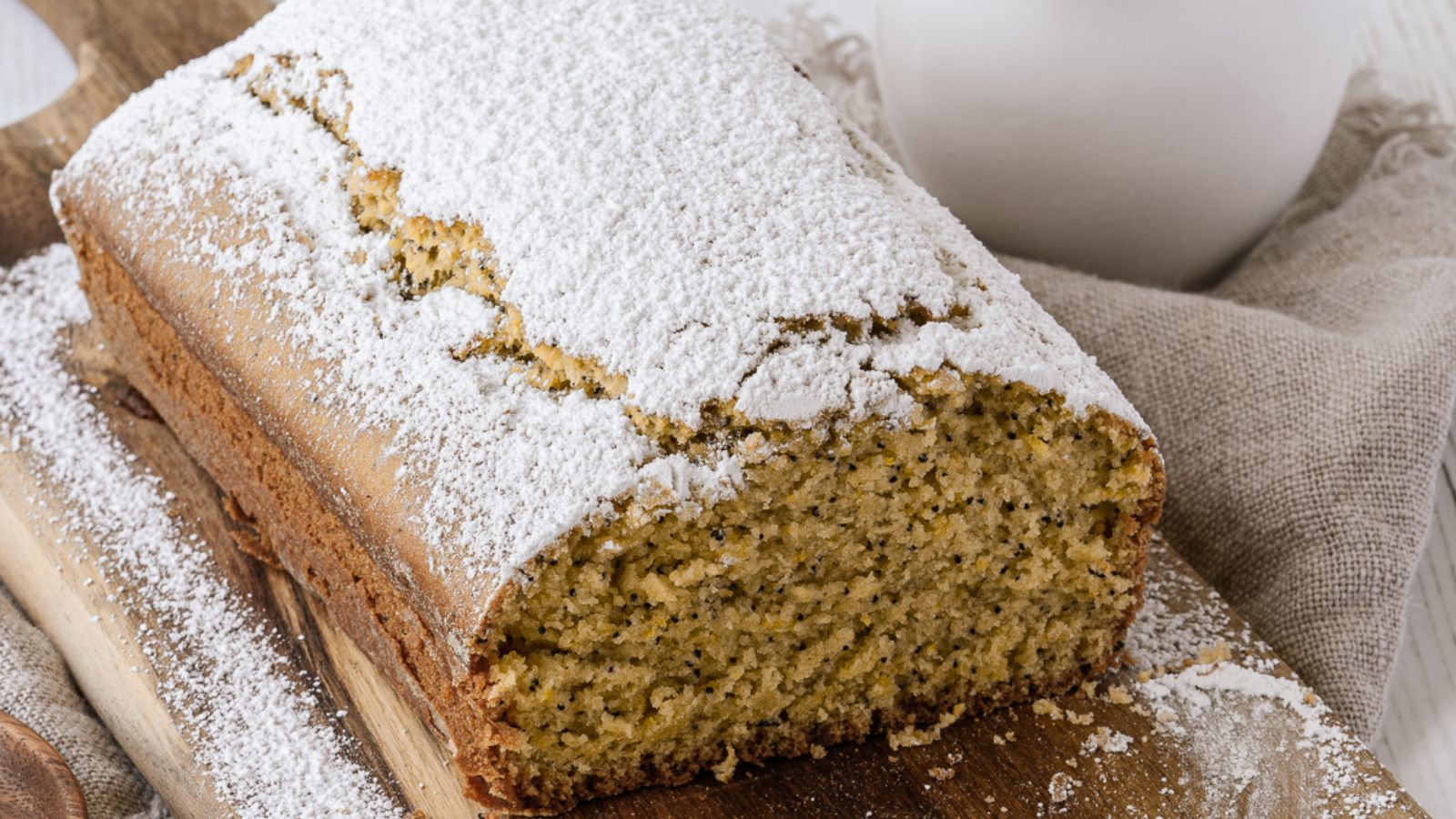 A loaf of lemon poppy seed bread dusted with powdered sugar sits on a wooden board with a beige cloth beside it.