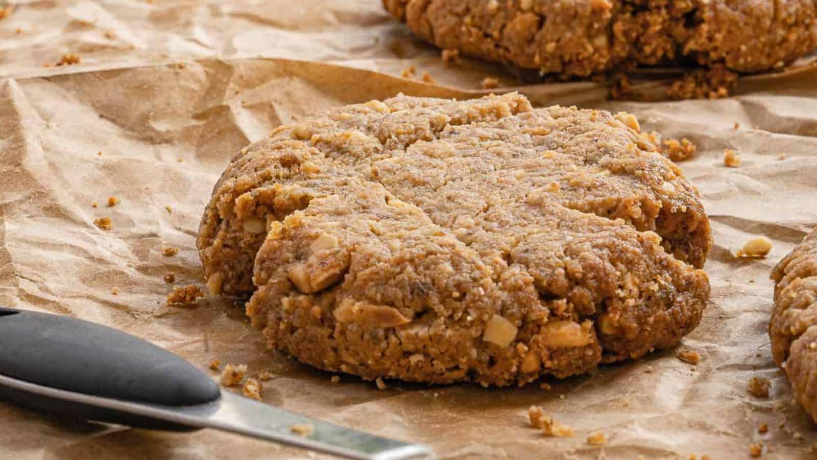 A close-up of a large, chunky peanut butter cookie with visible peanut pieces on brown parchment paper, next to a metal spatula with a black handle.