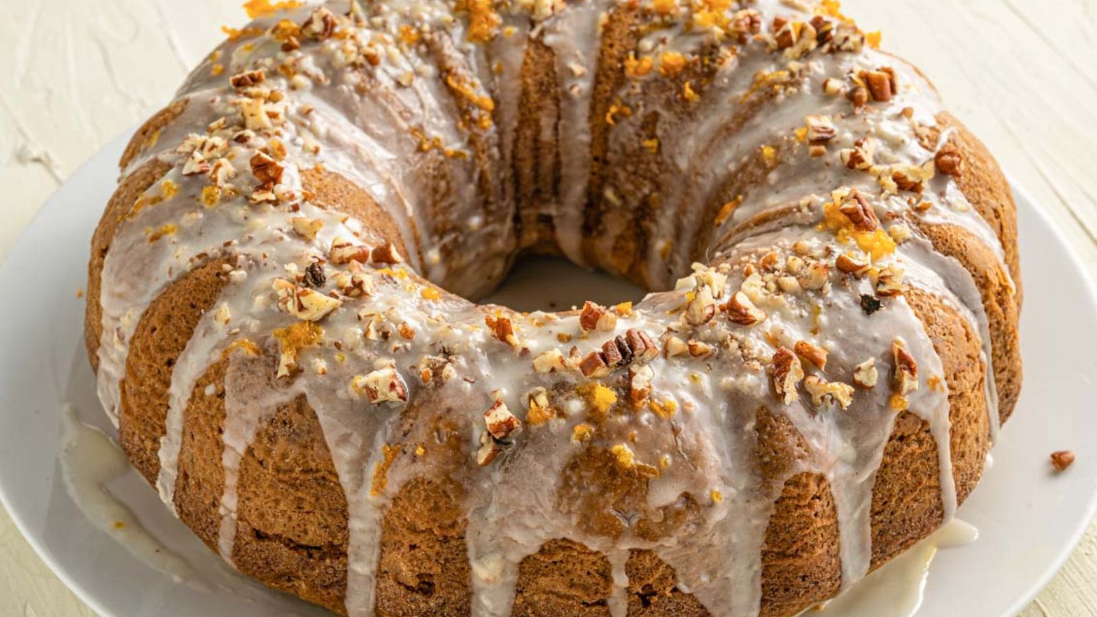 A bundt cake with white icing drizzled on top and sprinkled with chopped nuts, displayed on a white plate.