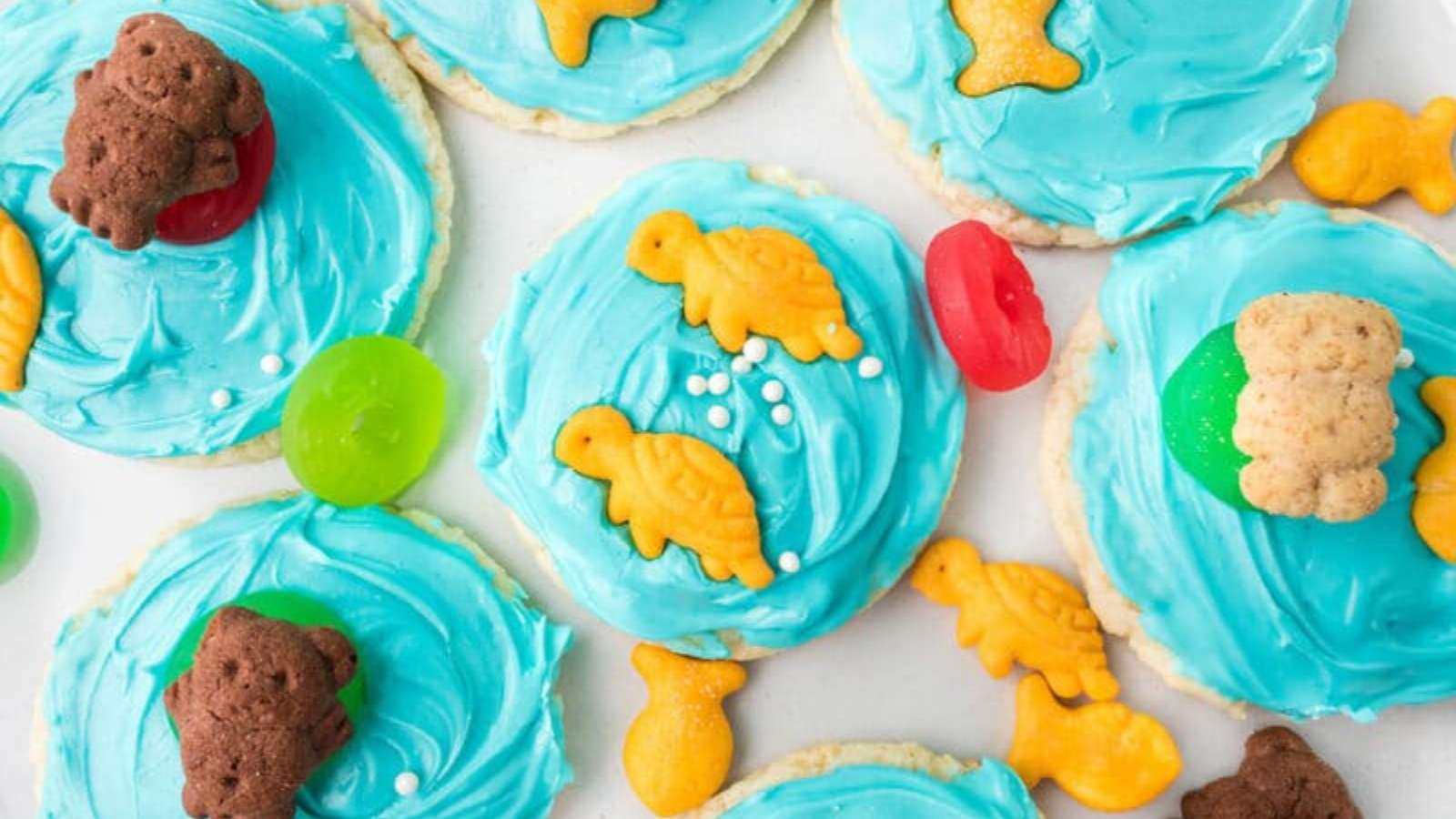Sugar cookies with blue frosting are topped with animal-shaped crackers, colorful gummy candies, and white sprinkles, arranged on a white surface.