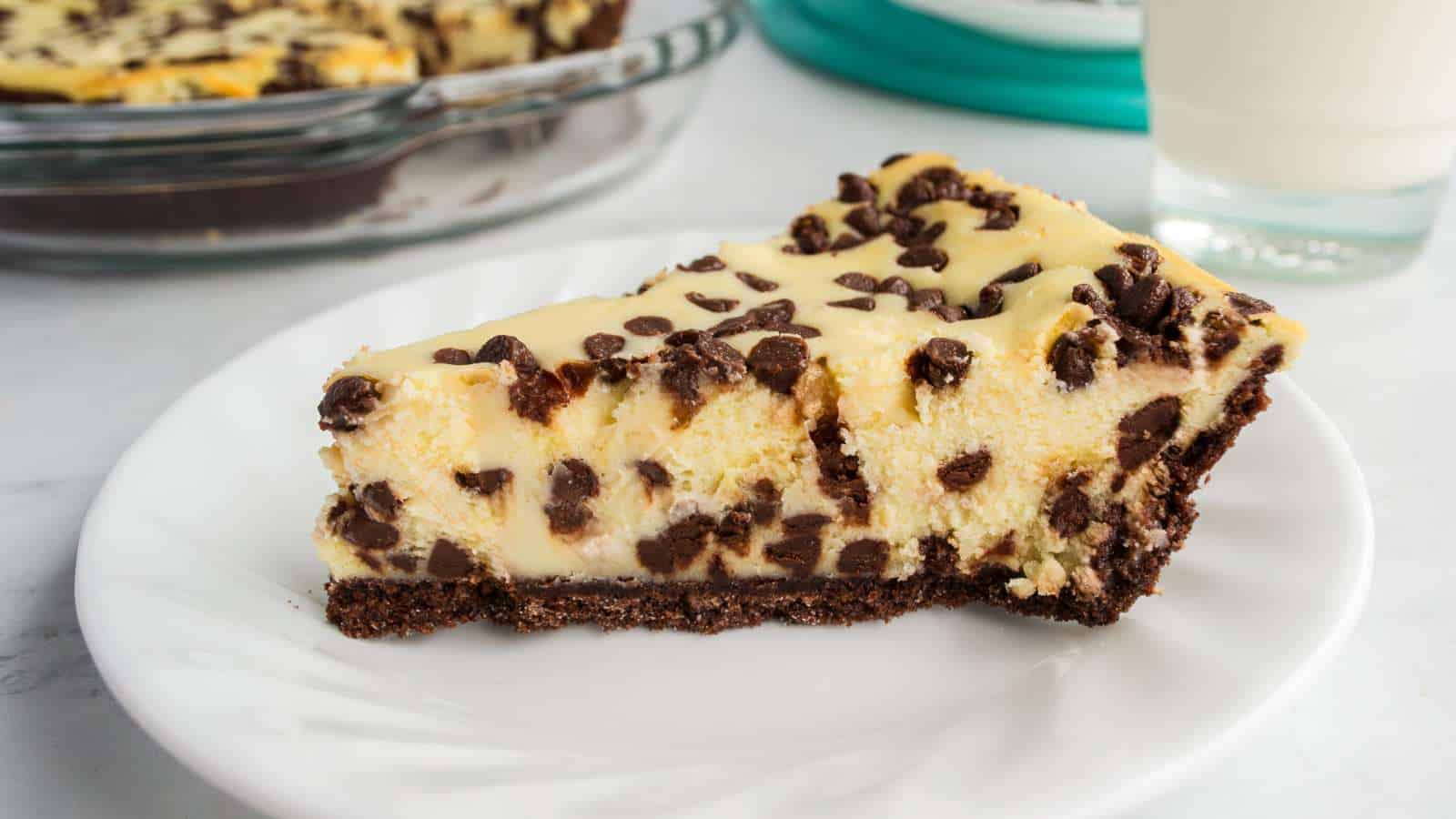 A slice of chocolate chip cheesecake with a chocolate crust on a white plate, with a glass of milk and a baking dish in the background.