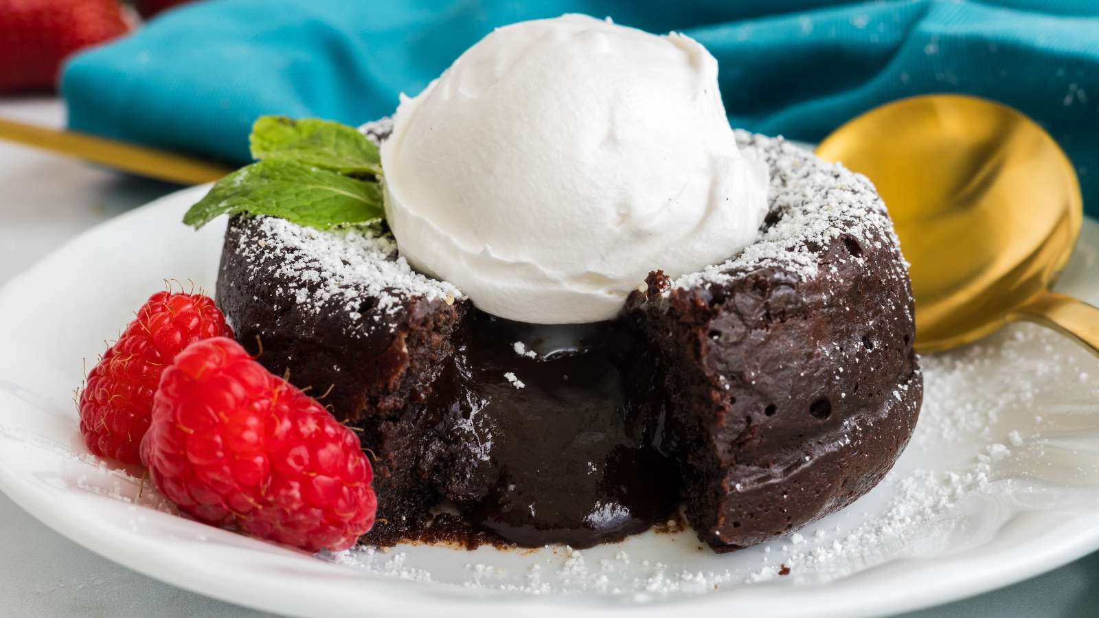 A chocolate lava cake with a molten center is topped with a dollop of whipped cream, mint leaf, raspberries, and powdered sugar on a white plate with a gold spoon.