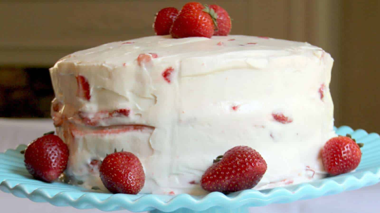 A round layer cake with white frosting and strawberries inside and on top, displayed on a blue cake stand.