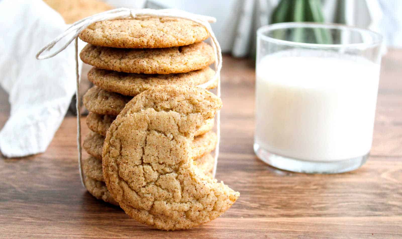 A stack of snickerdoodle cookies, with one cookie missing a bite, sits beside a glass of milk on a wooden surface.