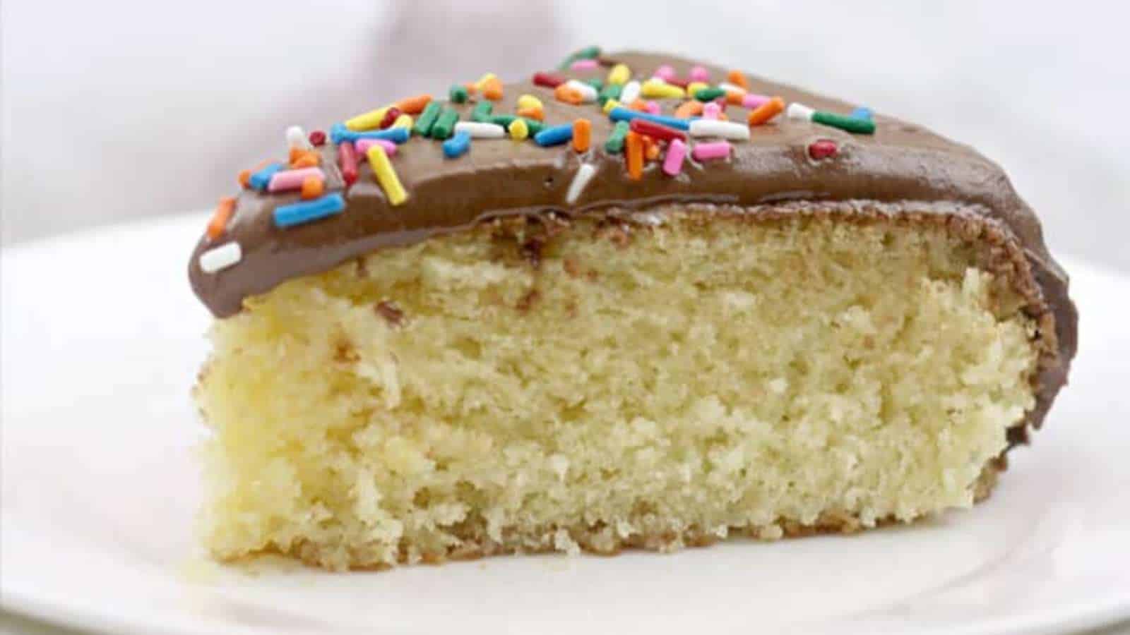 A slice of yellow cake with chocolate frosting and colorful sprinkles on top, served on a white plate.